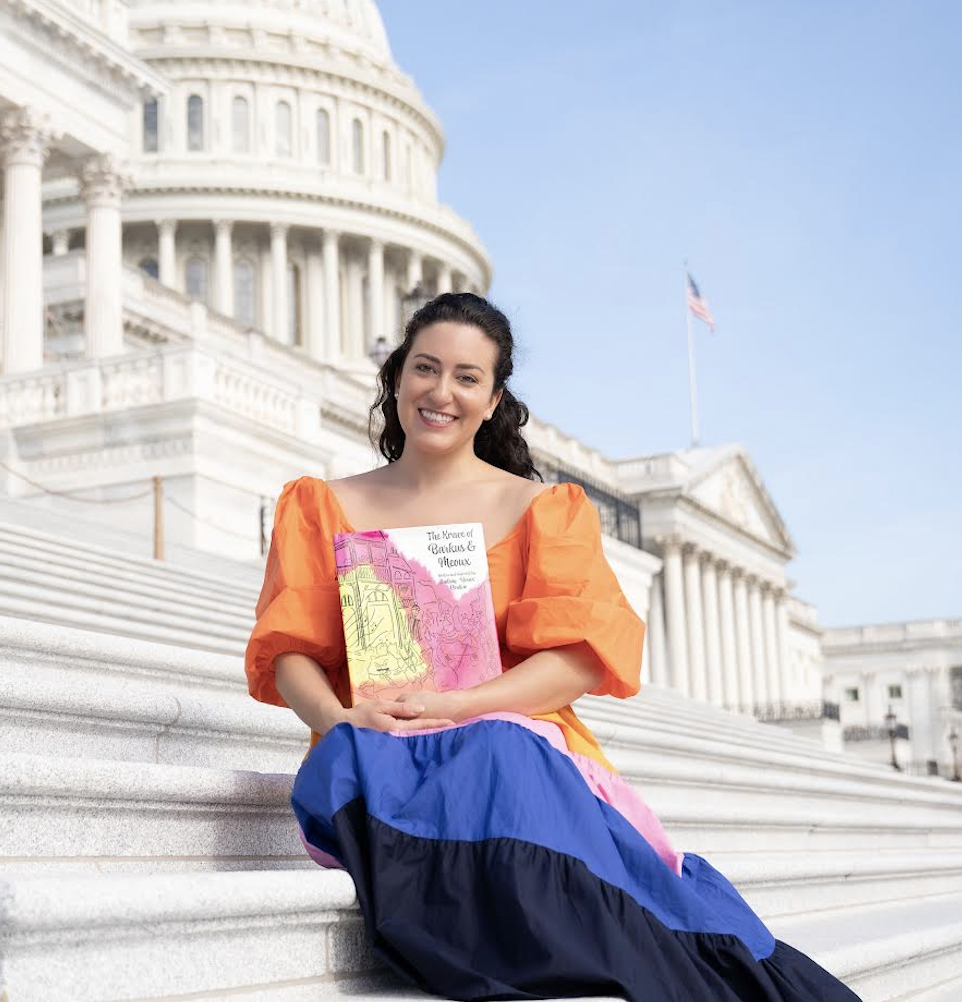 Photo of a woman with long dark hair sitting on the steps of a government building with the American flag waving in the background.