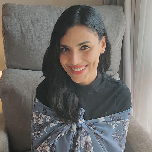 Woman sitting on a grey recliner smiling at the camera. She has medium length, wavy, black hair with a side part and dark eyes. She is wearing a black turtle neck top and has a blue scarf wrapped around her body with white flowers. 