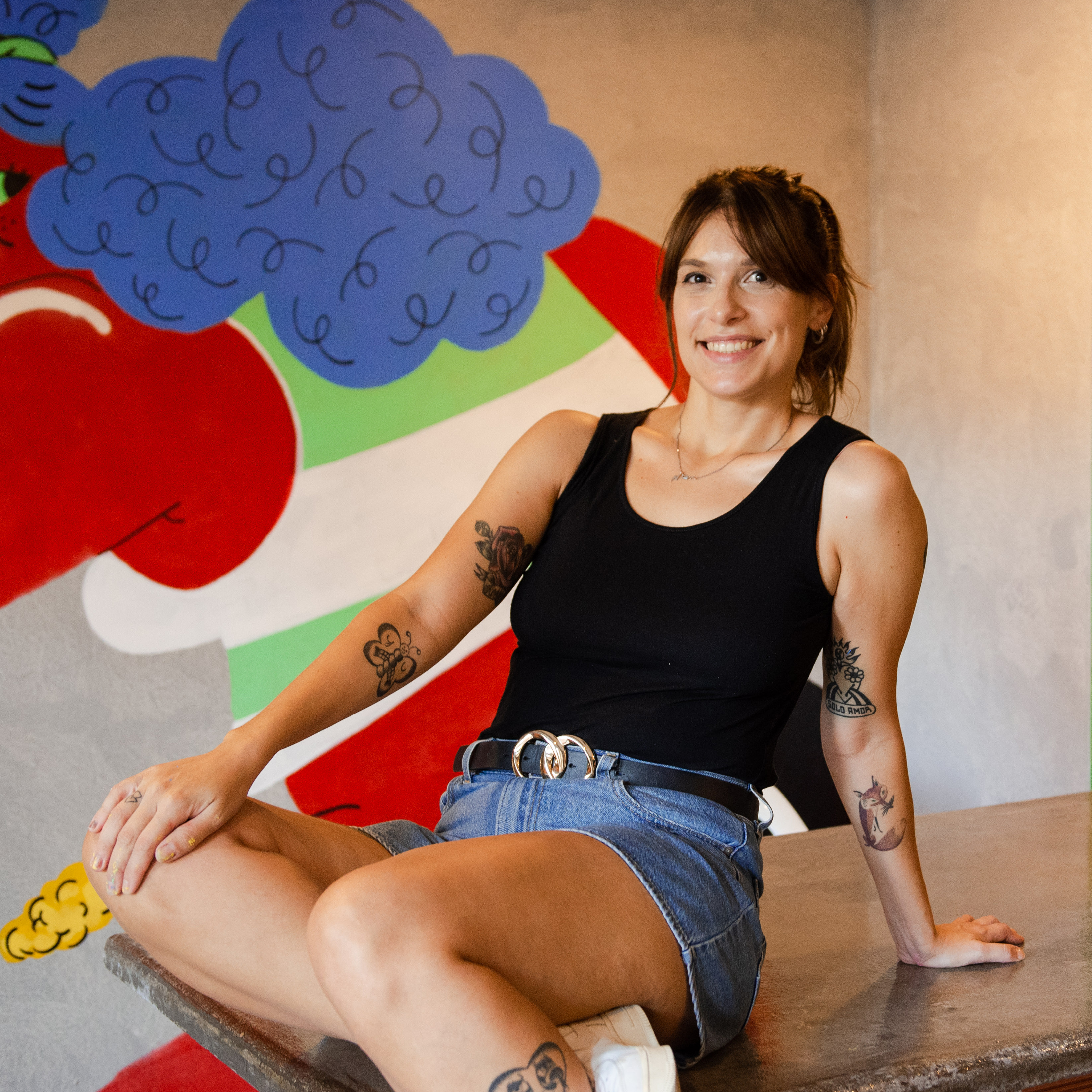 Photograph of a woman sitting, leaning backwards and smiling. She has tan skin, brown, pulled back hair, and is wearing a black t-shirt with jean shorts