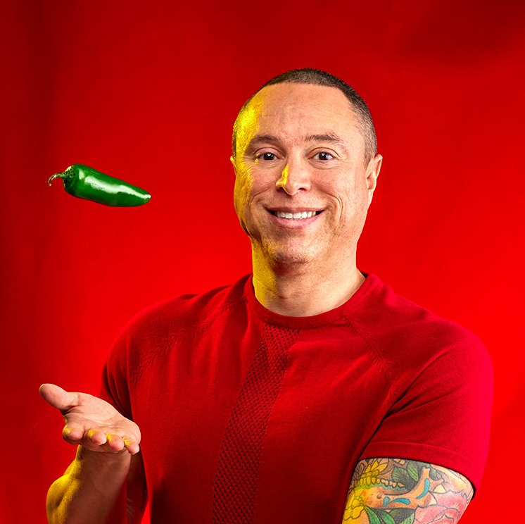 Photograph of a Hispanic man wearing a red shirt catching a jalepeño with his hand. 