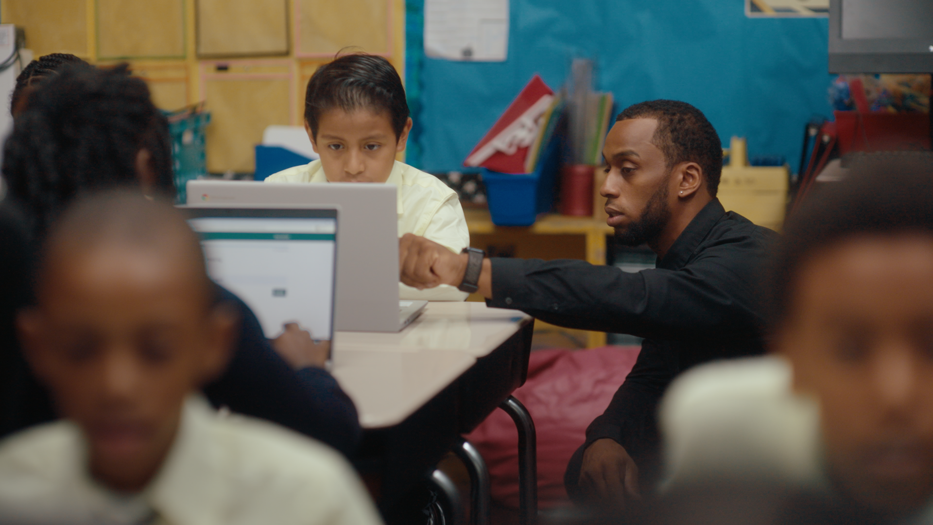 A teacher kneels by his student’s desk, hand pointing to the screen as his student focuses on the computer in front of him.