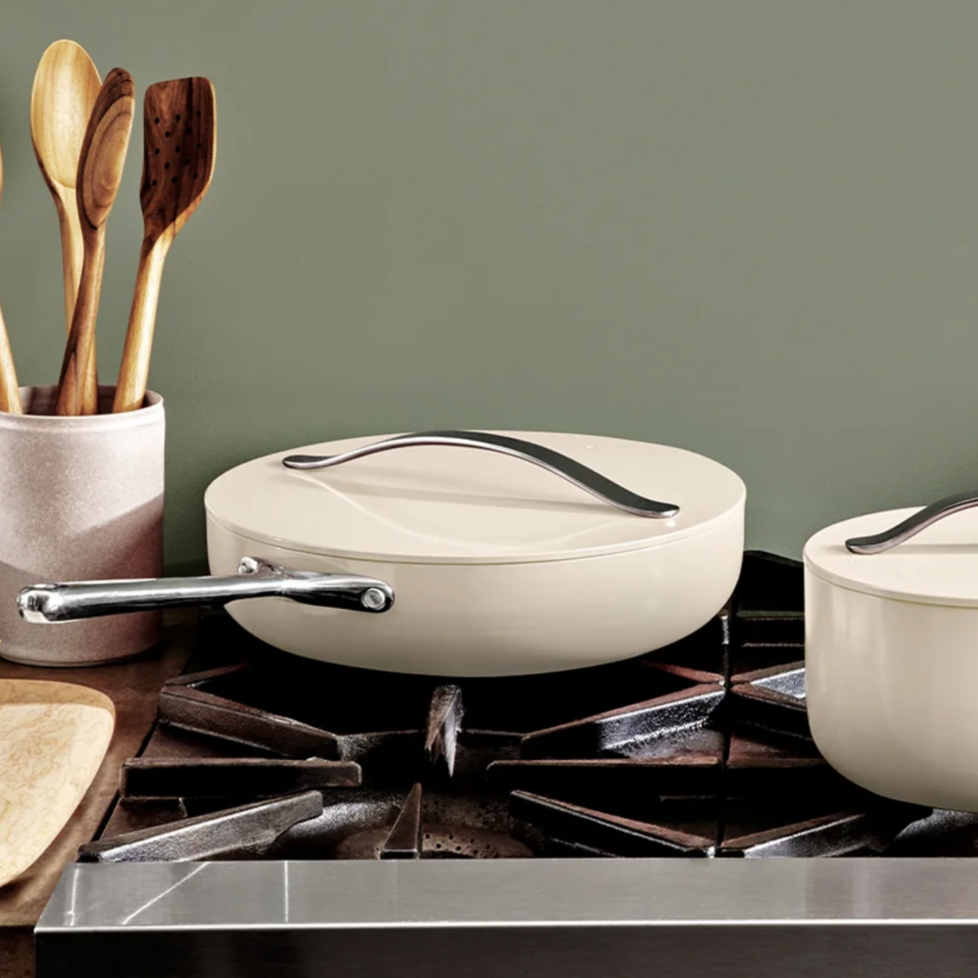 A Caraway pan on a stovetop next to a counter with wooden stirring utensils displayed on the left-hand side.