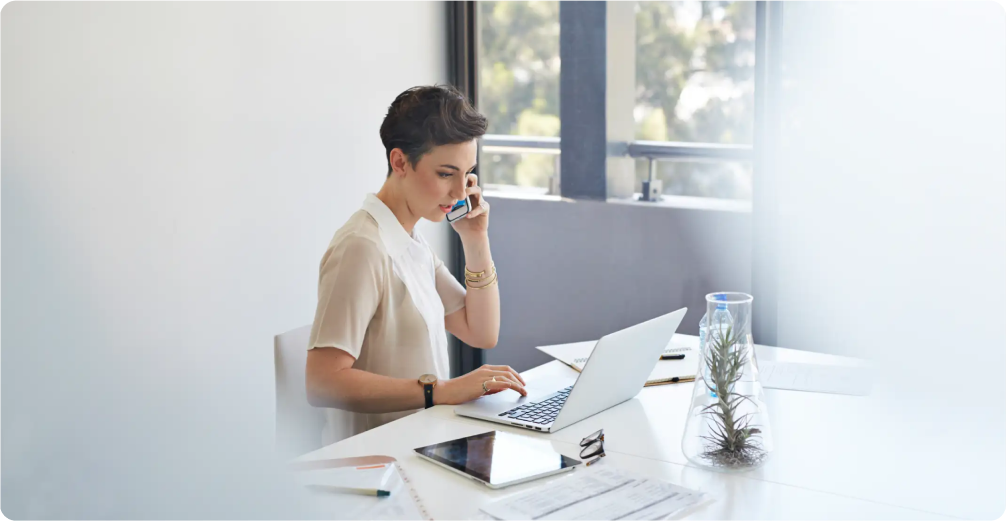 A short haired woman sits at a desk and talks on a smartphone with a laptop and tablet in front of her.