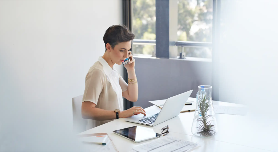 A short haired woman sits at a desk and talks on a smartphone with a laptop and tablet in front of her.