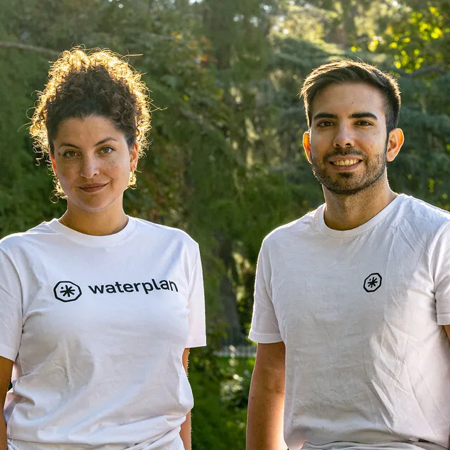 A group of four smiling individuals in white "Waterplan" t-shirts standing outdoors with trees in the background.