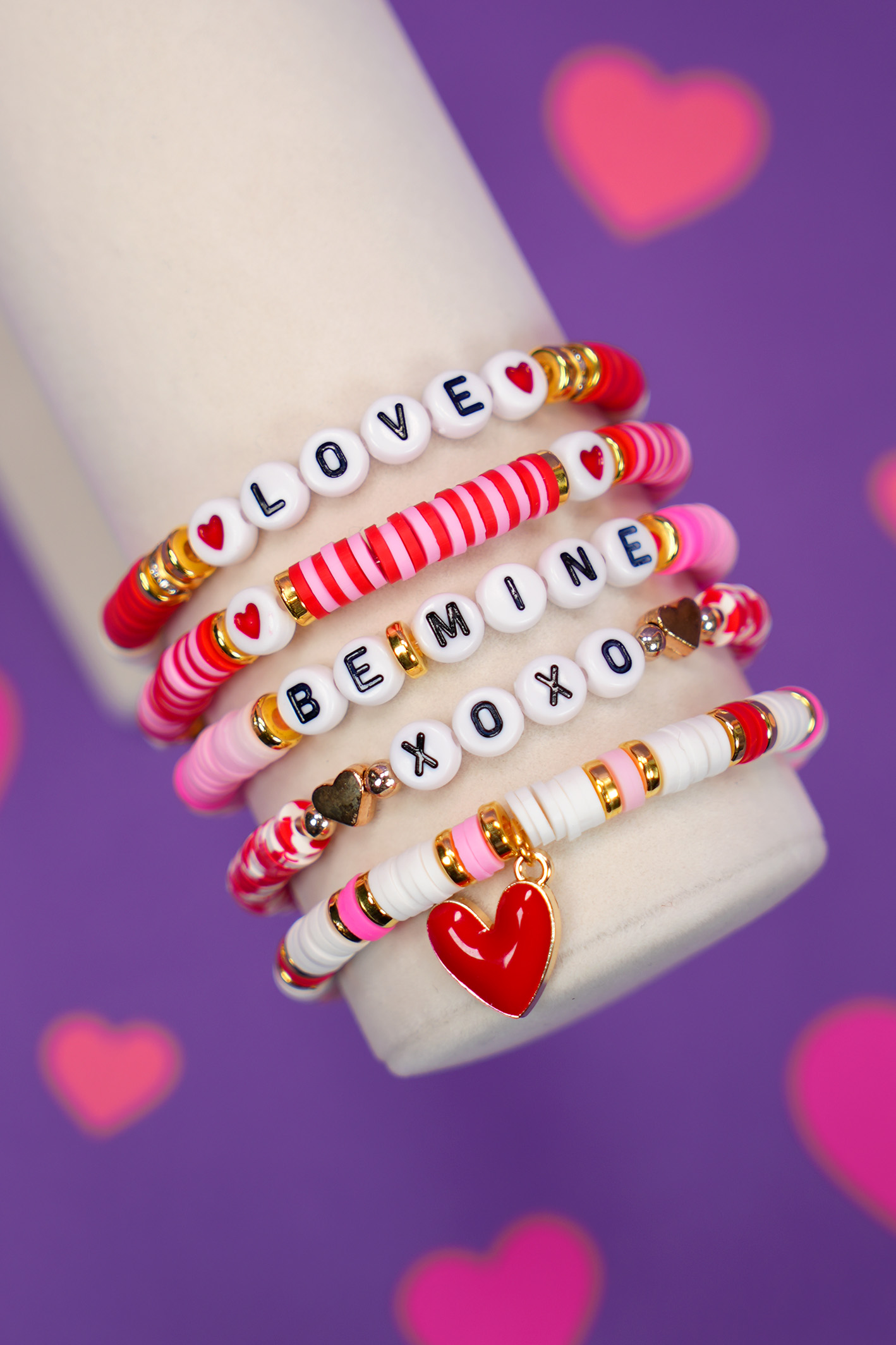 Close up of Valentine's Day bracelet set with red, pink, and white beads on display arm on purple background with pink hearts