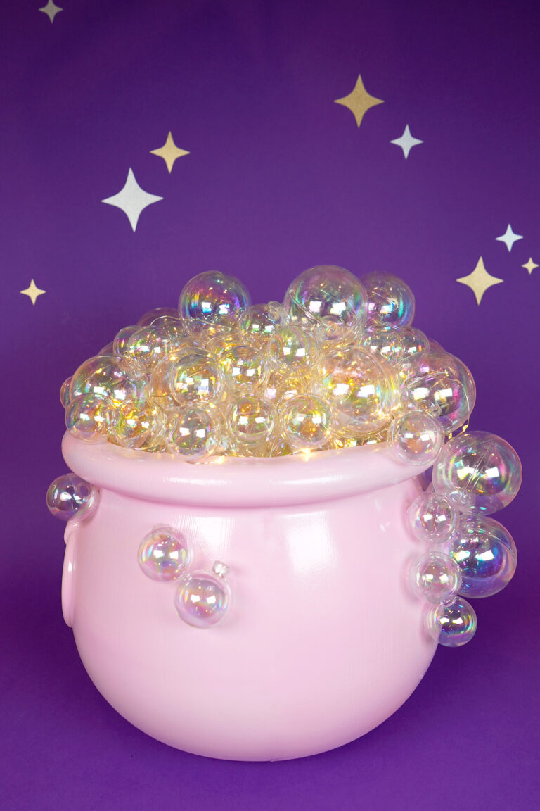 Pink plastic Halloween witch's cauldron with glowing iridescent "bubbles" against a dark purple background with gold and silver stars