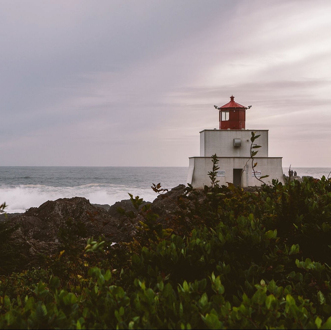 A short white lighthouse with a red top sits on the right side of the image o a rocky shorline with waves breaking in the background.