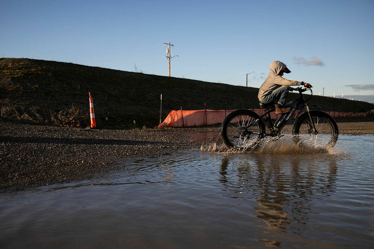 The Nosov family rides their bikes through a large puddle that formed next to the large piles of fill dirt at the Port of Everett on Monday, Dec. 9, 2024 in Everett, Washington. (Olivia Vanni / The Herald)