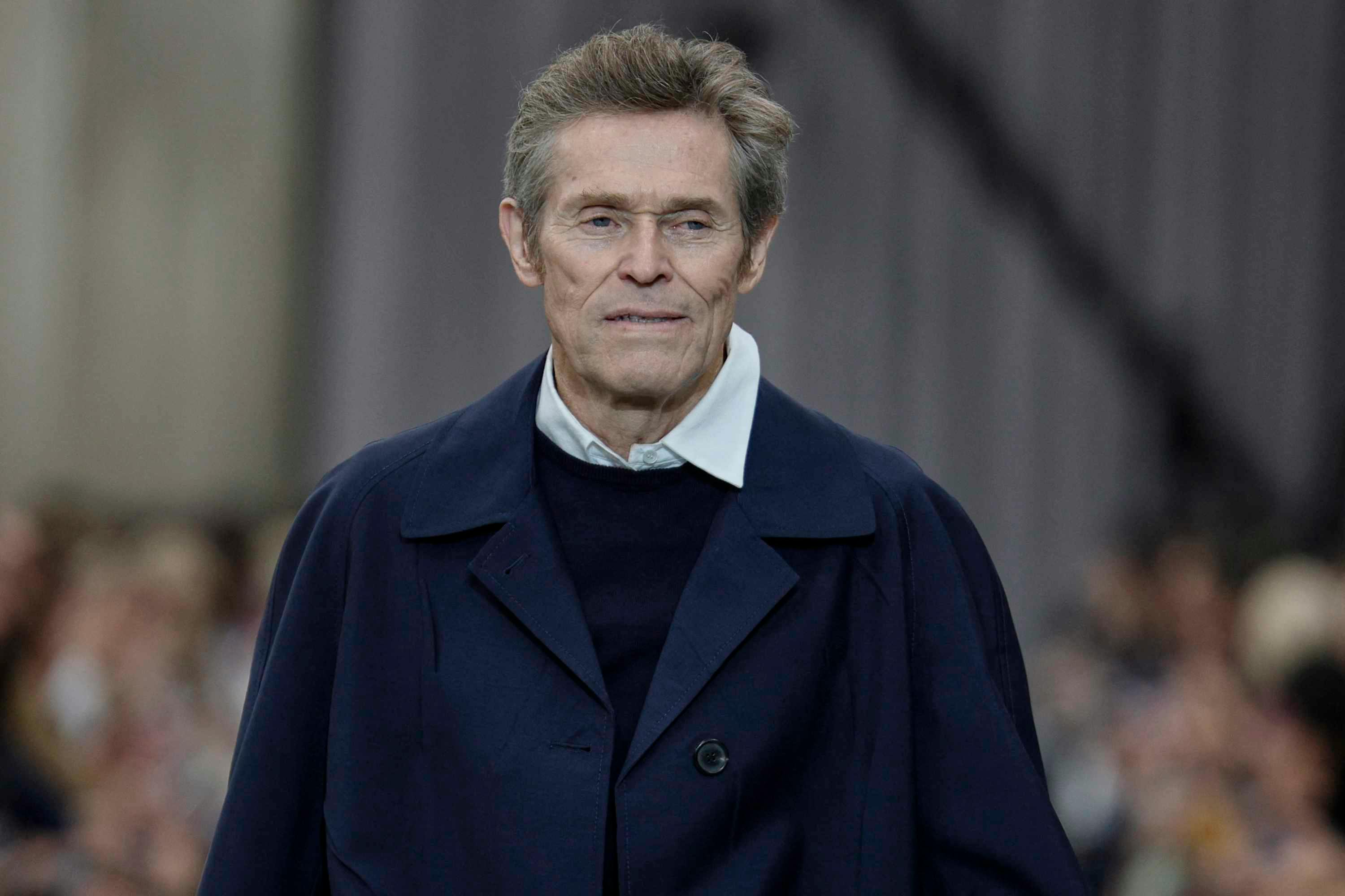 Willem Dafoe wears a navy miu miu coat, sweater and white shirt at the spring summer 2025 runway show