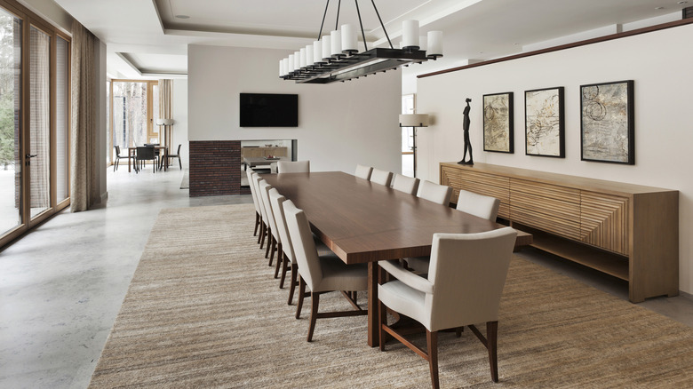 A large, modern dining room in neutral colors. The long wood table is flanked by beige chairs, and a sideboard is topped with modern art.