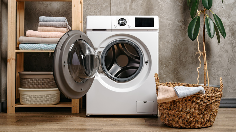 A washing machine with the door open in a modern laundry room. There is a shelf with folded towels, a plant, and a basket filled with laundry next to the machine.