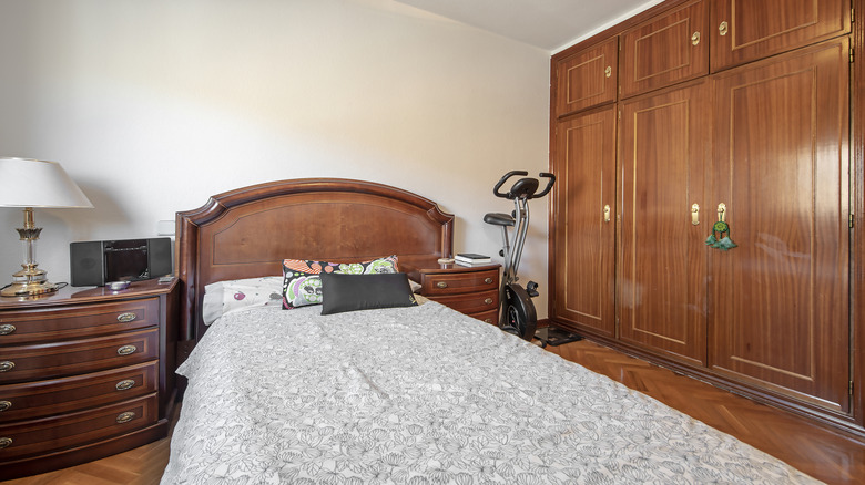 A cluttered bedroom with parquet floor, large wooden built-in xloset, and matching wooden bed frame and nightstands. The room has bare walls painted a light beige, and there is an exercise bike in the corner.