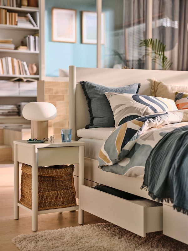 A bedroom with an off-white TONSTAD bedside table beside an off-white TONSTAD bed covered in MYGGLASVINGE multicolour bed linen.