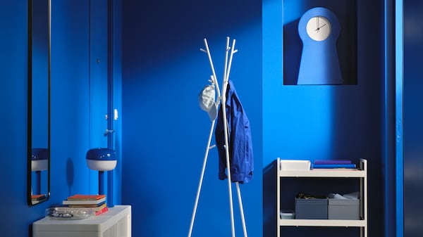 A blue hallway with an EKRAR hat/coat stand in the centre holding a coat and baseball cap, beside a white NISSAFORS trolley.