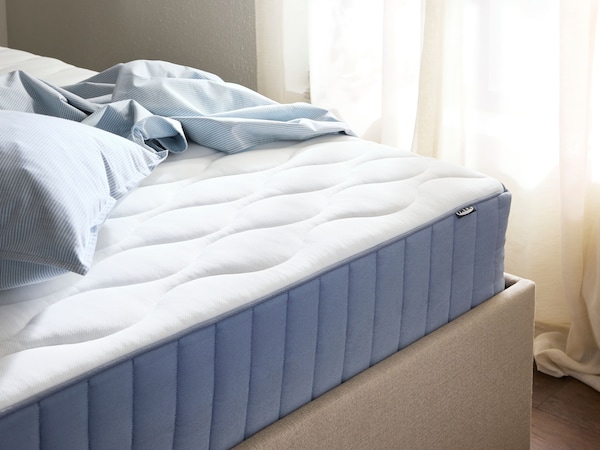 Close-up of beige bed frame with blue and white mattress, light blue sheets in a pile on top of it and a window behind.