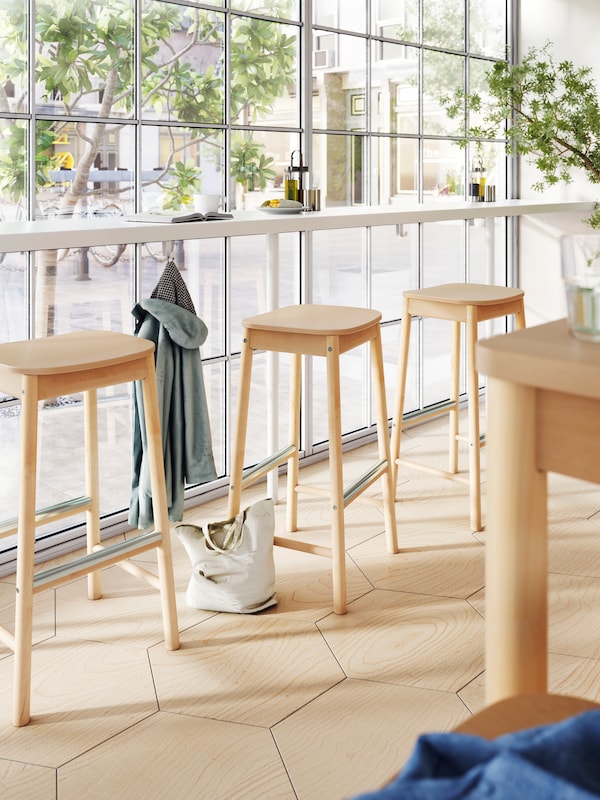 A long bar with a coat on a hook underneath and birch bar stools by a floor-to-ceiling window of a restaurant.