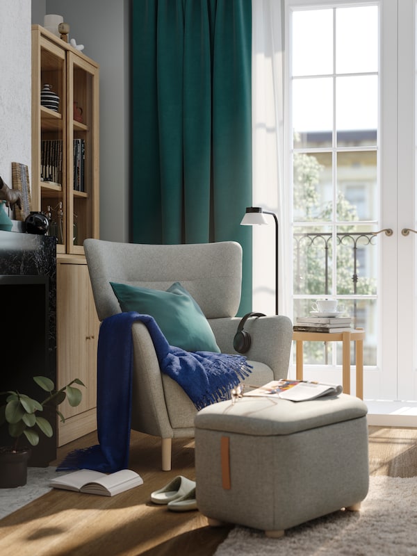 A living room with a sun-dappled beige-grey OSKARSHAMN wing chair and footstool with storage stand next to French windows.