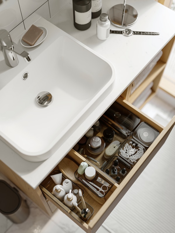 A drawer in a bathroom washstand is pulled out and inside are grey transparent VISSLAÅN boxes containing bathroom products.