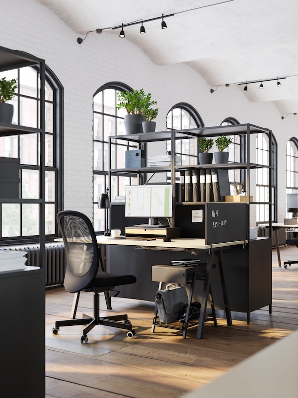 A TROTTEN desk and black office chair by open shelves with a privacy board at one side of the desk and a storage unit below.