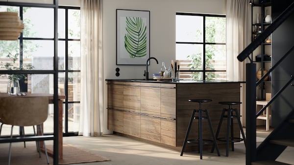 A kitchen island with STENSTA dark brown ash veneer fronts, complemented by bar stools and a large plant print on the wall.