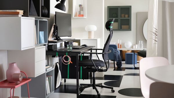 A side view of a PC gaming setup with a black UTVISNING gaming desk with shelf and a dark grey chair in a white passageway.