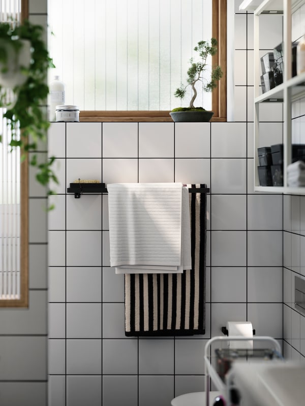 A solid coloured white towel and a black and white striped one, hanging neatly on a towel rail in a white tiles bathroom.