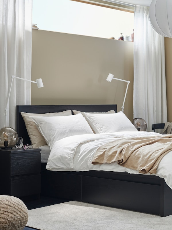 A beige, modern-style bedroom with a black-brown MALM bed with storage, and a bedside, black-brown MALM two-drawer chest.