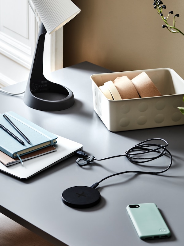 A desk with a black LIVBOJ wireless charger and some other objects on it: a smartphone, a laptop, notebooks and a desk lamp.