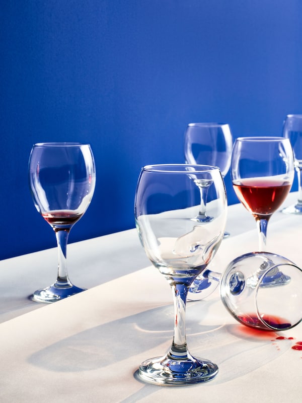 Several ÄNKEBLOMSTER wine glasses against a bright blue background. One of them is tipped over, a red liquid spilling out.