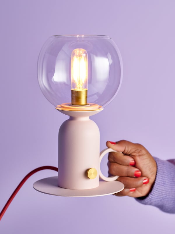 A hand holding a lit, dimmable ÅSKMULLER table lamp in light pink/red, against a solid lilac wall.