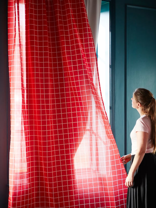 A woman holds red curtains and looks out as the sunlight comes in through the window.