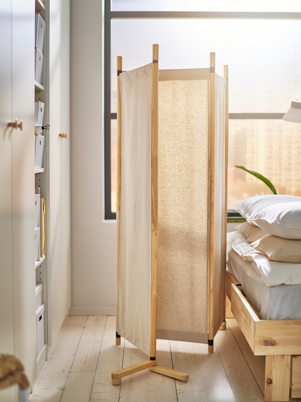 GLAMBERGET room divider in untreated wood and beige textile, standing between bed and wardrobe. Big window behind bed.