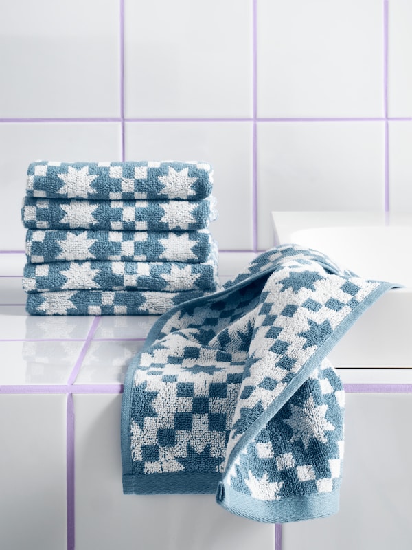 Some PILGULDMAL guest towels in dark grey-blue/white are folded on top of each other; one is unfolded on a tiled surface.