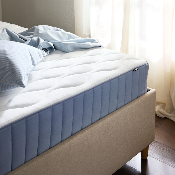 Close-up of beige bed frame with blue and white mattress, light blue sheets in a pile on top of it and a window behind.