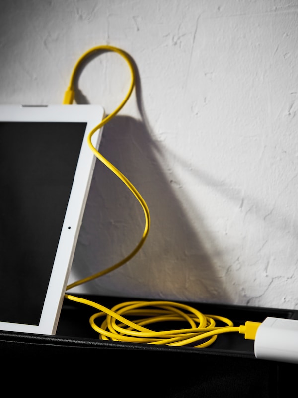 A yellow SITTBRUNN charging cable that’s wound up into a loose coil attaching a white battery pack to a white tablet.