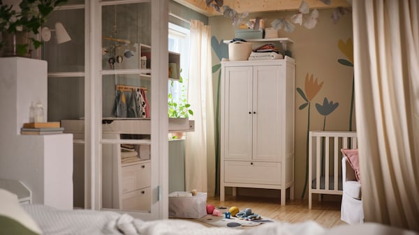 A white SUNDVIK wardrobe against a wall with a flower mural on it in a baby’s bedroom with a white SUNDVIK cot beside it.