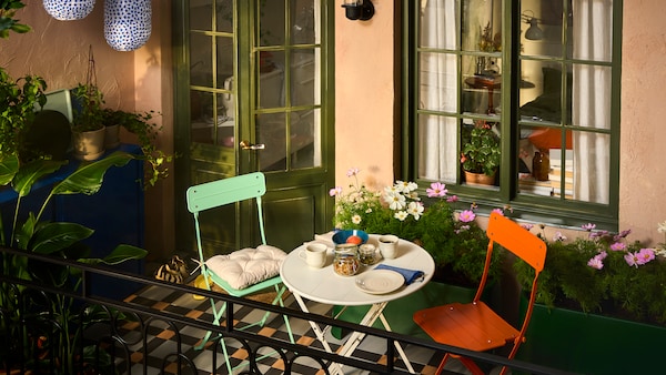 A balcony with SUNDSÖ outdoor furniture in playful colours surrounded by plants, flowers, lamps and cabinets for storage.