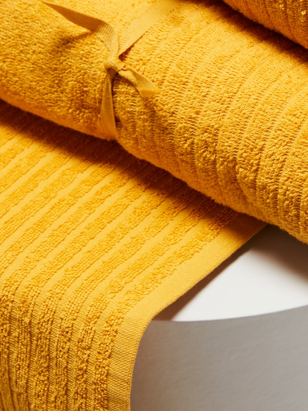 A closeup of a rolled-up yellow VÅGSJÖN towel on top of another VÅGSJÖN towel that is spread out over a white surface.