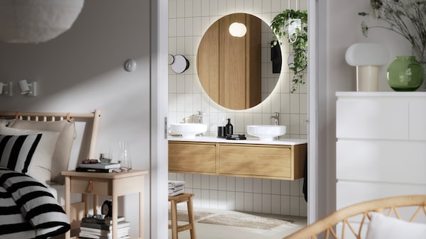 A modern bathroom with a round illuminated mirror, twin sinks, and wooden cabinetry, visible through an open door.