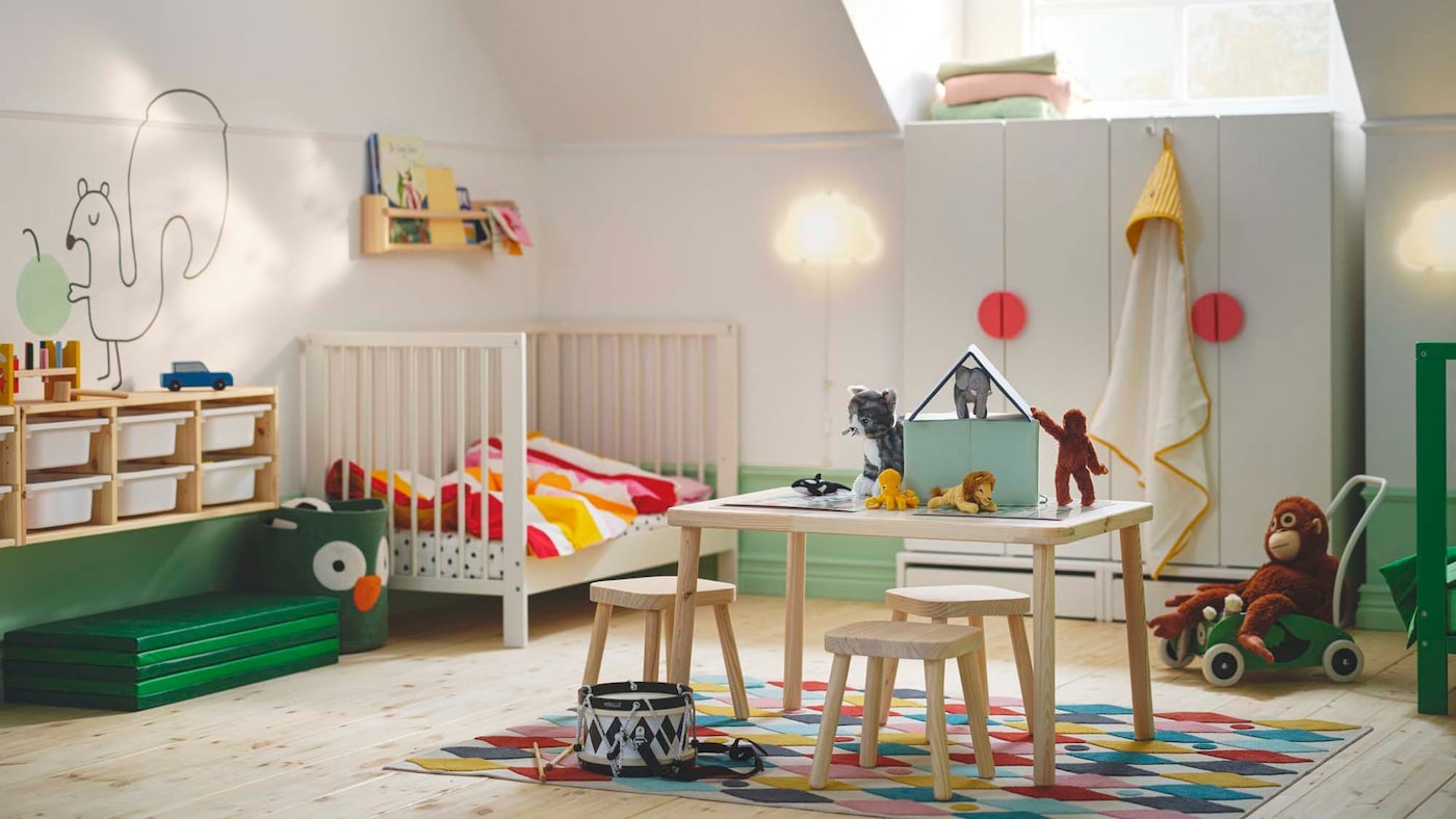 A todder bedroom in happy colours with a table and chairs in the centre, a white cot in the left-hand corner and TROFAST storage on the wall.