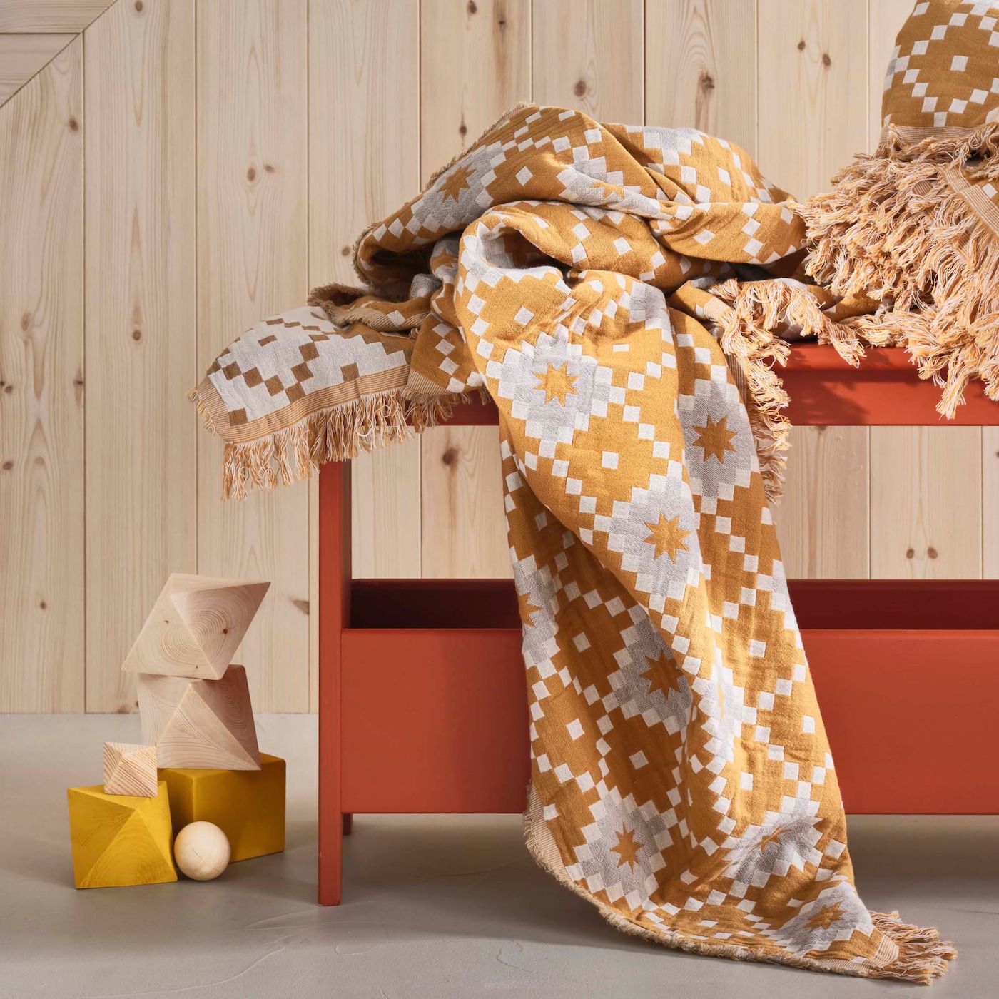 A yellow and cream SVARTFRYLE throw is piled on a terracotta coloured bench A corner drapes down onto the floor. In the background is a wood wall.