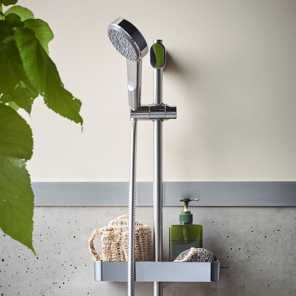 Chrome-plated, BROGRUND riser rail with handshower kit, mounted on a wall with a tray that holds a sponge and soap dispenser.