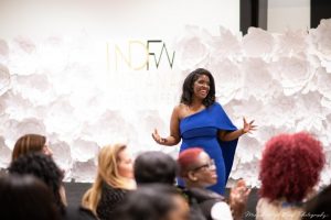 Women wearing a off the shoulder blue dress at Indiana Fashion Week in front of a group of people