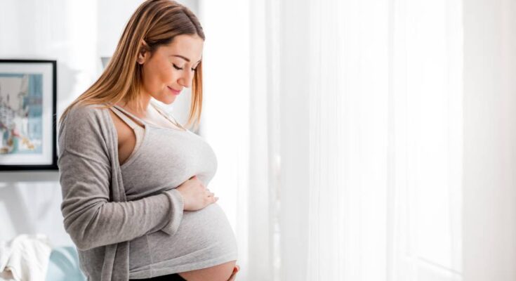 A pregnant woman in a gray tank top and cardigan standing near a sunny window, smiling as she looks down at her baby bump.