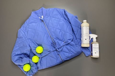 Auf einem grauen Untergrund liegt eine blaue Steppjacke mit drei gelben Tennisbällen. Neben der Jacke stehen zwei Flaschen mit Reinigungsmitteln.