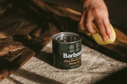 Eine Person trägt mit einem gelben Schwamm Wachs auf ein braunes Kleidungsstück auf. Eine Dose mit der Aufschrift „Barbour Wax Thornproof Dressing“ steht in der Nähe auf einer mit Stoff bedeckten Oberfläche. Die Szene ist warm beleuchtet, und die Schatten lassen auf eine natürliche Lichtquelle schließen.