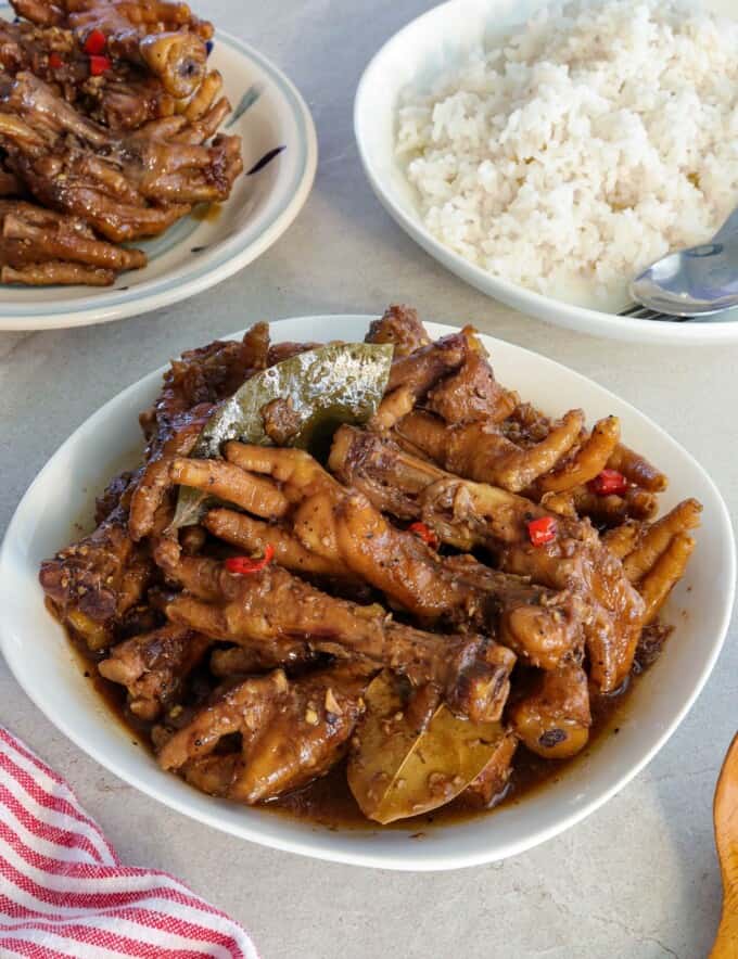 adobong paa ng manok in a white bowl with a plate of steamed rice in the background.