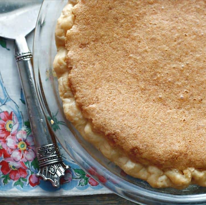 The upper crust of a chess pie in a glass pie plate with a silver serving handle and flowered napkin