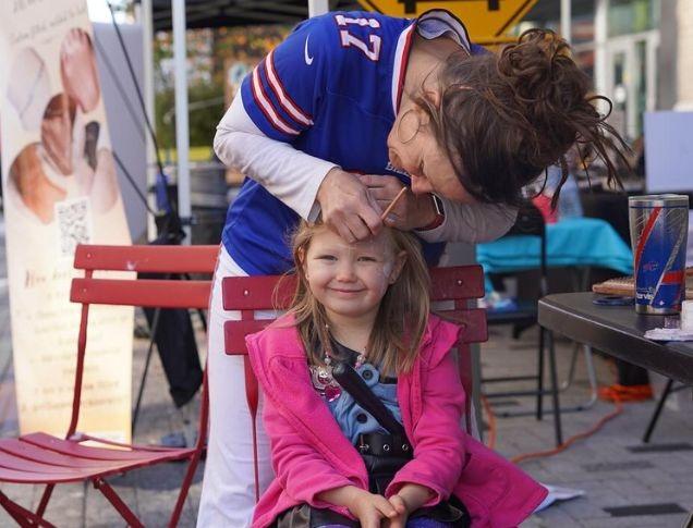 A smiling young girl in a pink jacket sits on a red chair as a woman, dressed in a blue sports jersey, painting her face.